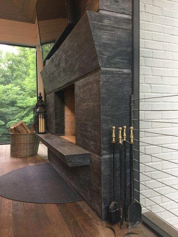 Modern dark stone fireplace with angled hood in contemporary interior space with brick wall, wooden flooring, and large window overlooking trees